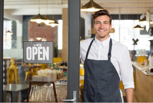 A smiling business owner standing at the entrance of a shop with an 'Open' sign, representing Small Business Development.
